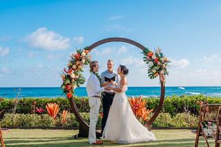 A couple exchanges vows under a floral arch by the ocean, with a celebrant guiding the ceremony amidst a beautiful blue sky.