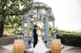 A couple poses romantically under an elegant gazebo surrounded by lush vineyards, celebrating their love in a picturesque setting.