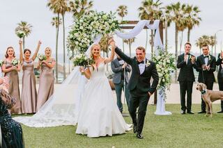 A joyful couple celebrates their wedding, surrounded by bridal party and guests, with palm trees in the backdrop. Love and cheer fill the air.