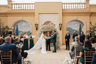 A couple exchanges vows during a wedding ceremony, surrounded by guests and a bridal party in elegant attire, with a floral backdrop.