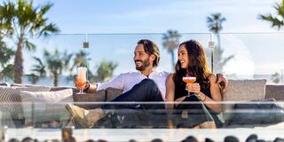 A cheerful couple enjoys cocktails on a sunny terrace, surrounded by palm trees and a beautiful blue sky, radiating happiness and relaxation.