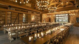 A spacious rustic dining area with wooden beams, large tables set with candles, and warm lighting creates an inviting atmosphere.