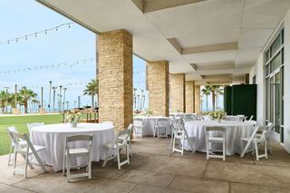 A stylish outdoor venue features round tables with white tablecloths and elegant floral arrangements, surrounded by palm trees and sea views.