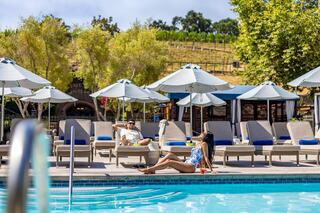 A relaxed scene by a pool, featuring two people lounging on chairs under umbrellas, surrounded by lush greenery and a vineyard backdrop.