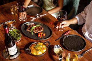 A beautifully set dining table features two steaks, garnished tomatoes, green asparagus, creamy side dish, and glasses of red wine.