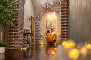 A romantic scene features a couple in vibrant attire seated together in a softly lit, brick-lined corridor adorned with candles.