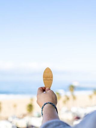 A hand holds a wooden surfboard-shaped object against a backdrop of ocean waves and palm trees under a clear blue sky.