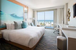 A modern hotel room with a large bed, light colors, and a stunning ocean view through glass doors. Perfect for relaxation.