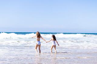 A woman and a girl hold hands, wading into the ocean, enjoying a sunny day at the beach with gentle waves and clear skies.