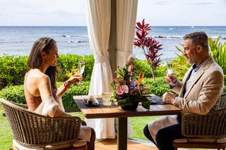 A couple enjoys drinks at an elegant table, overlooking the ocean, surrounded by vibrant flowers and lush greenery.