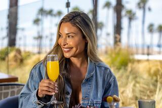 A joyful woman with long hair smiles while holding a mimosa, surrounded by palm trees and a sunny outdoor setting.