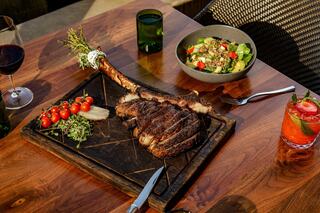A succulent tomahawk steak rests on a wooden board, accompanied by cherry tomatoes, a fresh salad, and drinks in a stylish setting.