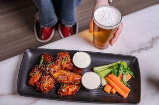 Crispy chicken wings drizzled with sauce, served with two dips, celery, and carrots, alongside a cold beer in hand.