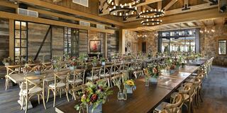 A spacious dining area features long wooden tables adorned with flower arrangements, rustic decor, and large windows for natural light.