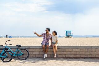 A couple sits on a sand wall, chatting and enjoying the beach view, with a bike nearby and a lifeguard tower in the distance.