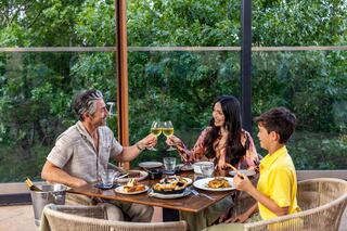A family shares a joyful meal, raising glasses in a toast amid a vibrant green backdrop, enjoying delicious dishes together.