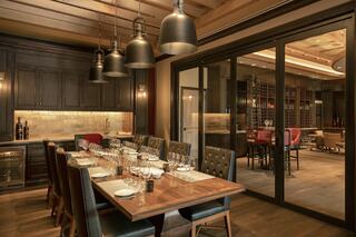 A stylish dining room features a large wooden table, elegant table settings, and modern lighting, with a wine cellar visible through glass.