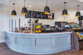 A modern café counter features a curved design, glass barriers, coffee machines, snacks, fruits, and stylish pendant lighting overhead.