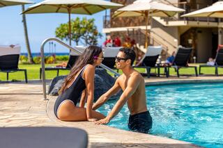 A couple enjoys a sunny day by the pool, playfully engaging with each other while surrounded by lush scenery and lounge chairs.