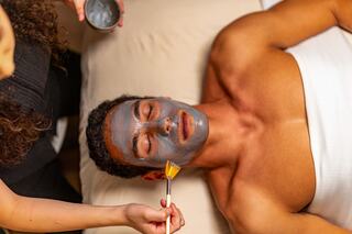 A relaxed individual receives a facial treatment, with a grey mask being applied by a person using a brush. Soft lighting creates a calm atmosphere.