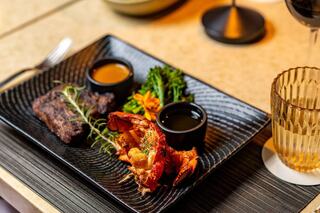 A gourmet plate featuring succulent lobster, grilled meat, vibrant broccoli, and sauces, elegantly arranged on a textured black platter.