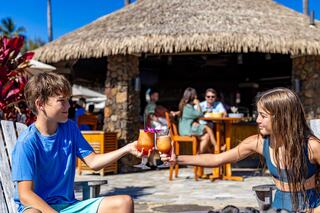 Two young children toast with colorful drinks outside a tropical bar, surrounded by a sunny atmosphere and people enjoying their time.