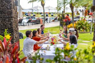 A lively outdoor gathering features friends toasting with drinks at a beautifully set table, surrounded by lush greenery and a scenic backdrop.