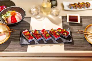 A beautifully arranged sushi platter features vibrant pieces topped with colorful garnishes, accompanied by bowls and chopsticks on a textured mat.