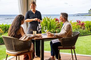 A couple enjoys a scenic meal by the ocean while a waitress serves them, surrounded by lush tropical greenery.