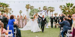 A joyous wedding celebration outdoors, with the couple cheering as guests applaud amidst palm trees and elegant decorations.