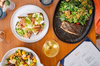 A vibrant spread featuring a fresh salad, a savory fish dish, and a toast topped with greens and cured meat, complemented by a glass of wine.