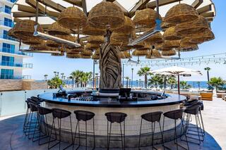 A stylish outdoor bar features a unique canopy of woven light fixtures, surrounded by palm trees and ocean views, inviting relaxation.