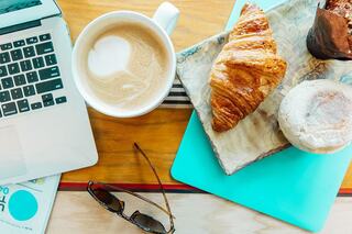 A laptop rests on a wooden table beside a cup of coffee, a croissant, and a dessert, with sunglasses and a magazine nearby.