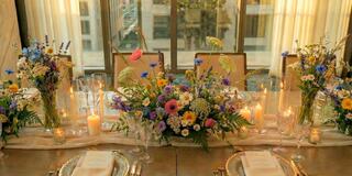 A beautifully arranged dining table features vibrant floral centerpieces, elegant glassware, and glowing candles, creating a warm, inviting atmosphere.