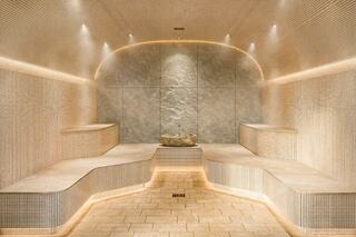 A serene steam room features softly illuminated, curved stone seating and a calming central fountain, creating a tranquil atmosphere.