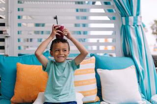 A young boy smiles brightly while holding up a drink in celebration, surrounded by colorful cushions in a cozy outdoor setting.