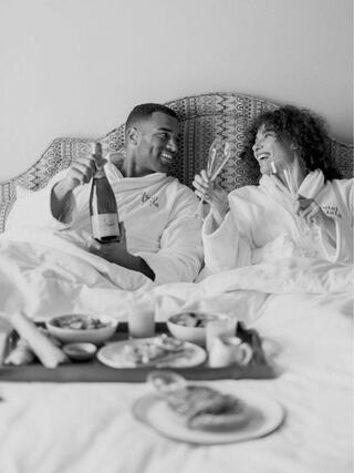 A couple enjoys a cozy breakfast in bed, wearing matching robes, sharing laughter and champagne, surrounded by delicious food.