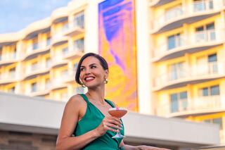A woman in a green dress smiles while holding a cocktail, with a vibrant building backdrop showcasing colorful lighting.