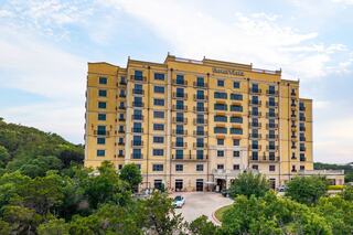 A modern hotel with a yellow facade and multiple balconies stands amidst lush greenery, offering a welcoming view against a clear sky.