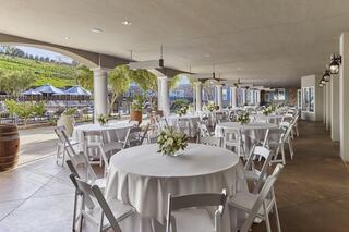 A serene outdoor venue featuring elegantly set round tables with white tablecloths and floral centerpieces, surrounded by greenery.