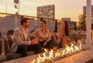 Three people relax on a rooftop during sunset, enjoying drinks by a fire bowl, with a stylish cityscape in the background.