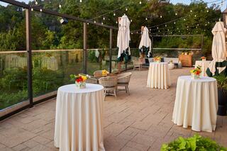 A cozy outdoor terrace features neatly arranged tables with floral centerpieces, elegant umbrellas, and string lights amid lush greenery.