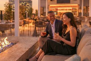 A couple enjoys drinks by a cozy fire on a rooftop, surrounded by elegant decor and a warm sunset ambiance.