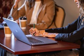 A person writes notes while seated at a table, with a laptop and coffee cups present, suggesting a productive meeting environment.