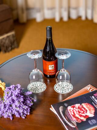 A bottle of red wine sits between two glasses on a wooden table, accompanied by lavender and a magazine, creating a cozy atmosphere.
