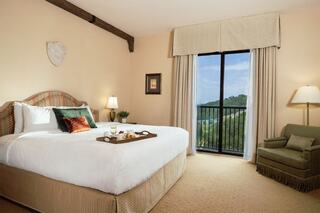 A cozy hotel room features a large bed, bedside lamps, a chair, and a balcony with scenic views, complemented by tasteful decor.