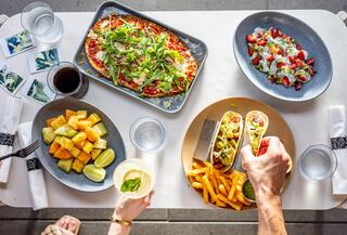 A vibrant spread showcases a pizza with greens, tacos with toppings, fresh fruit, and colorful sides, paired with drinks on a stylish table.