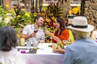 A joyful couple toasts at a lively outdoor gathering, surrounded by friends and vibrant tropical flowers, sharing happiness and good drinks.