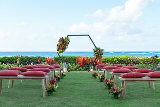 A beautiful outdoor wedding setup features wooden benches with red cushions, floral arrangements, and a serene ocean backdrop.
