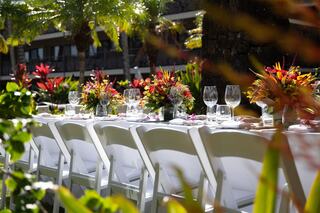 A beautifully set outdoor dining table adorned with vibrant floral centerpieces, surrounded by lush greenery and elegant glassware.